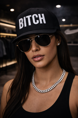 Person wearing a black cap with 'BITCH' and sunglasses in an indoor setting