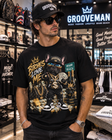 A man wearing a black Grooveman t-shirt with a detailed graphic of a French bulldog wearing sunglasses and a gold chain, standing inside a retail store.