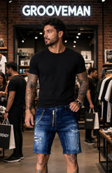 A man wearing a black crewneck t-shirt and distressed blue denim shorts with a silver zipper detail, standing inside a clothing store with 'GROOVEMAN' illuminated on the wall behind him.