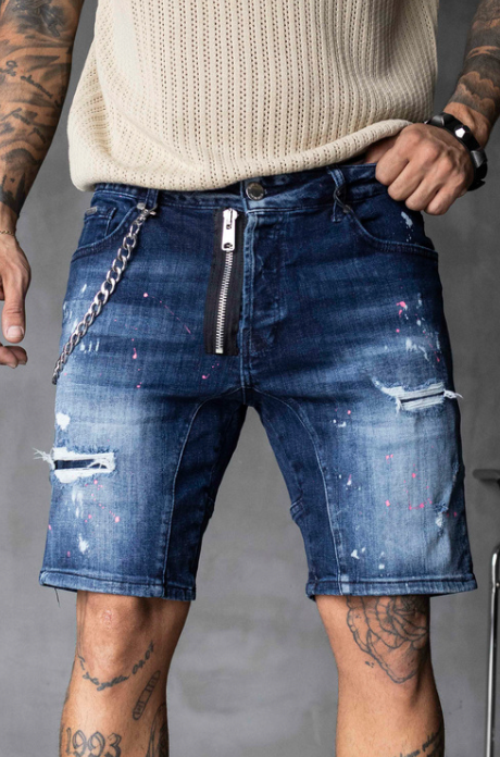 A close-up of a person wearing distressed blue denim shorts with a front zipper, chain detail, and paint splatter accents, paired with a cream knit sweater against a gray wall.