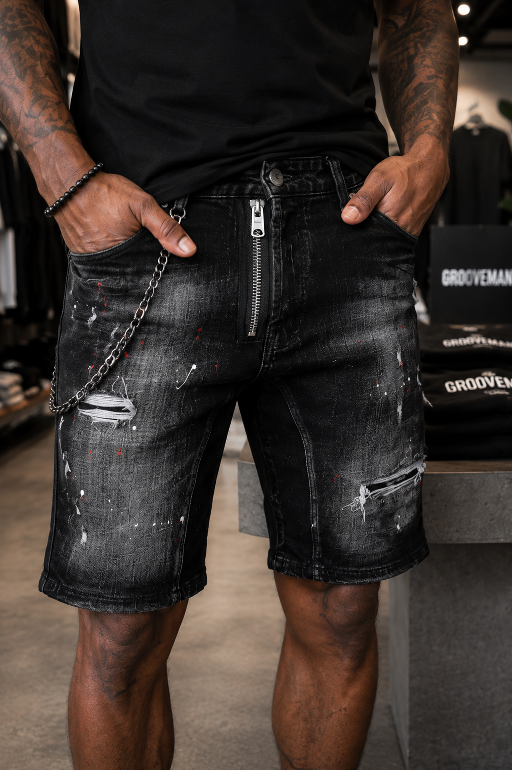 A close-up of a person wearing distressed black denim shorts with a front zipper, chain detail, and white paint splatter print, standing in a retail store.