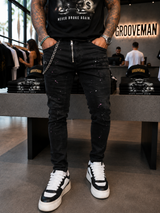 A person standing in a retail store wearing black distressed skinny jeans with a silver chain detail at the waistband, a black graphic t-shirt, and black-and-white high-top sneakers. The background shows a clothing store interior with other customers and Grooveman branding.