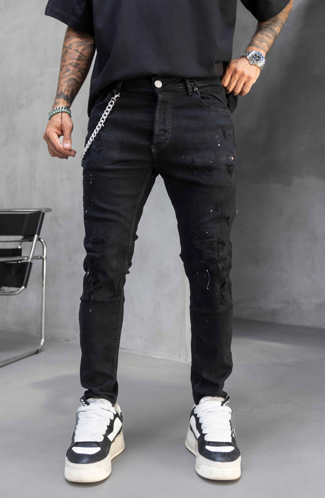 A person wearing distressed black skinny jeans with a silver chain detail at the pocket, paired with a black t-shirt and black-and-white sneakers, standing against a gray studio background.