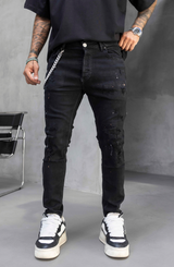 A person wearing distressed black skinny jeans with a silver chain detail at the pocket, paired with a black t-shirt and black-and-white sneakers, standing against a gray studio background.