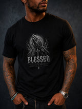 A man wearing a black graphic t-shirt with a detailed grayscale print of praying hands and the word 'BLESSED' on the front, set against a dark textured background.
