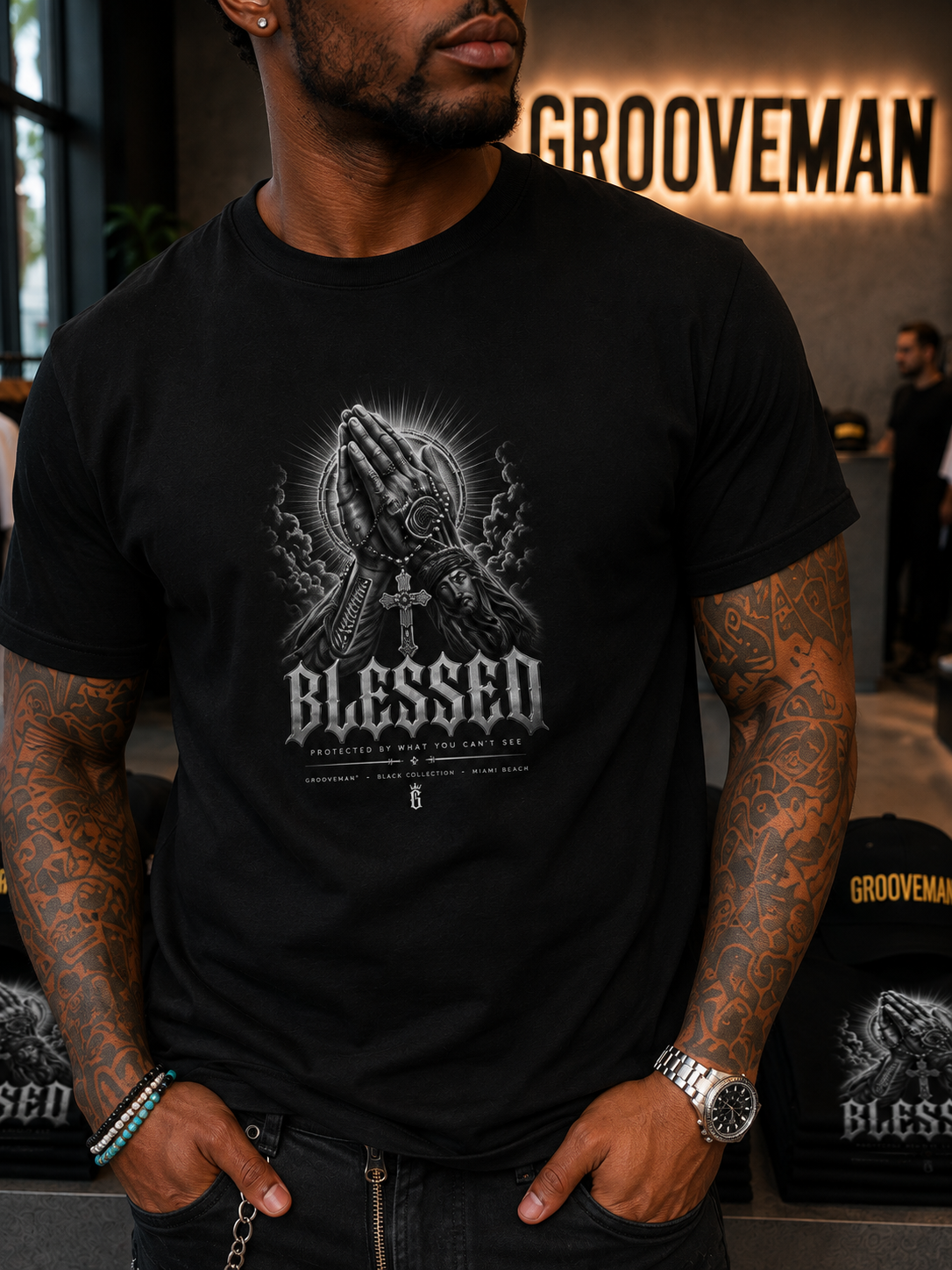 A man wearing a black crewneck t-shirt with a detailed grayscale graphic of praying hands and the word 'BLESSED' on the front, set in a gym environment with a Grooveman sign in the background.