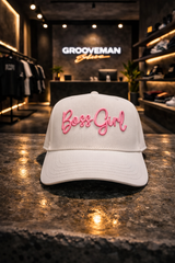 White cap with 'Boss Girl' in pink text on a reflective surface, with a store interior in the background.