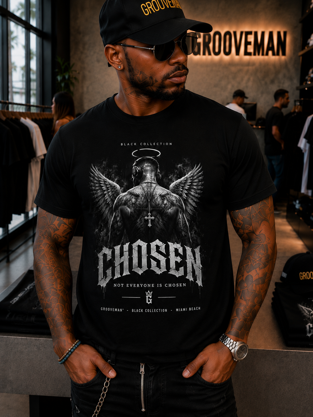 A man wearing a black t-shirt with a detailed white graphic of an angel and the word 'CHOSEN', standing in a clothing store with 'GROOVEMAN' branding on the wall behind him.
