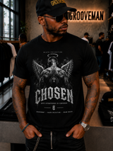 A man wearing a black t-shirt with a detailed white graphic of an angel and the word 'CHOSEN', standing in a clothing store with 'GROOVEMAN' branding on the wall behind him.