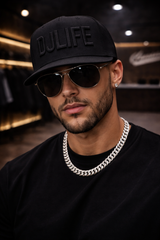 Man wearing a black cap with 'DJ LIFE' text, sunglasses, and a chain necklace in a dark indoor setting.