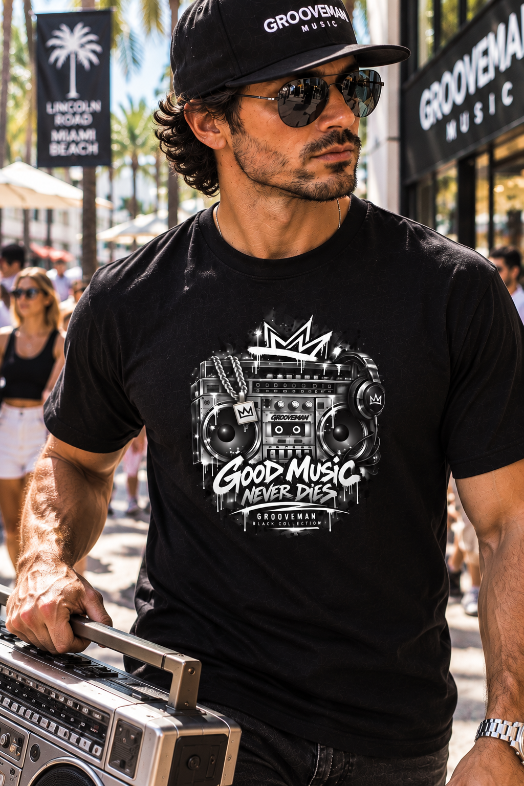 A man wearing a black Groovemain branded t-shirt with a white retro boombox graphic, sunglasses, and a matching cap, carrying a vintage boombox in a sunny outdoor street setting.