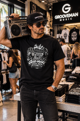 A man wearing a black Groovemusic t-shirt with a white retro boombox graphic, a black cap, and sunglasses, holding a vintage boombox over his shoulder inside a retail store.
