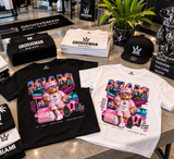 A retail display of Groovemusic Miami Beach branded apparel, featuring black and white t-shirts with a colorful graphic of a teddy bear in a pink outfit next to a pink car, along with matching black caps and folded shirts on a polished stone counter.