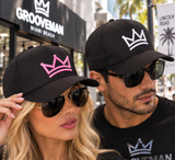 A man and a woman wearing black baseball caps with crown logos in white and pink, styled with sunglasses in an outdoor street setting.