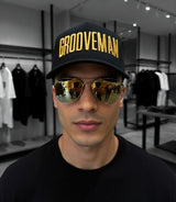 Man wearing a black cap with 'GROOVEMAN' and reflective sunglasses in a clothing store.