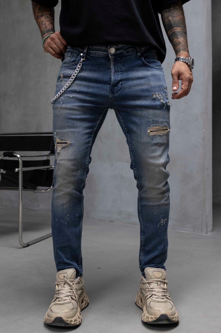 A person wearing distressed blue denim jeans with ripped details, a silver chain belt, a black t-shirt, and beige sneakers, standing in a studio setting.