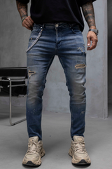 A person wearing distressed blue denim jeans with ripped details, a silver chain belt, a black t-shirt, and beige sneakers, standing in a studio setting.