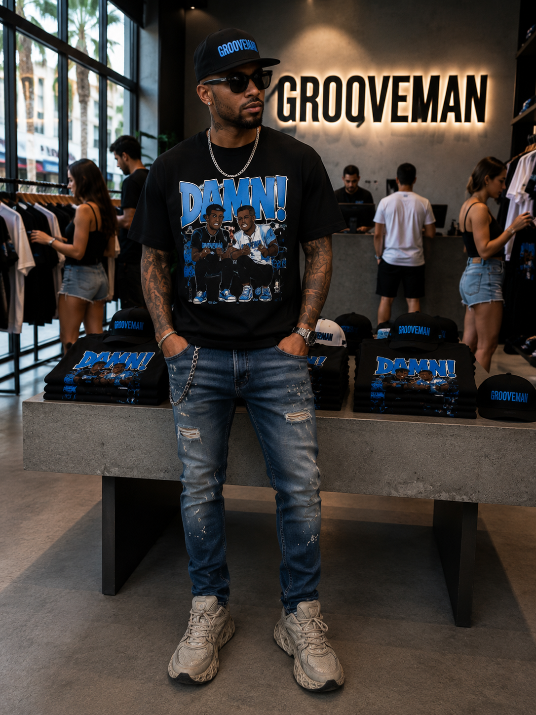 A man wearing a black Grooveman t-shirt with a blue graphic print, blue jeans, and a black cap stands in a retail store. The shirt features the word 'GROOVEMAN' on the back in blue and white, and the front has a blue 'GROOVEMAN' logo above a photo of a group.