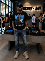 A man wearing a black Grooveman t-shirt with a blue graphic print, blue jeans, and a black cap stands in a retail store. The shirt features the word 'GROOVEMAN' on the back in blue and white, and the front has a blue 'GROOVEMAN' logo above a photo of a group.