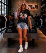A woman stands in a retail store wearing a black graphic T-shirt with a colorful '35 Years Miami' design, light-wash denim cutoff shorts, white high-top sneakers, and holding a pink drink. The background shows clothing racks, shelving, and a branded wall sign reading 'NOVEMAN'.