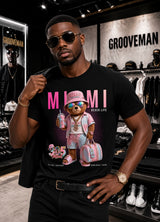 A man wearing a black Grooveman t-shirt with a colorful graphic of a teddy bear dressed in pink beachwear under the text 'MIAMI BEACH LIFE', standing inside a retail store.
