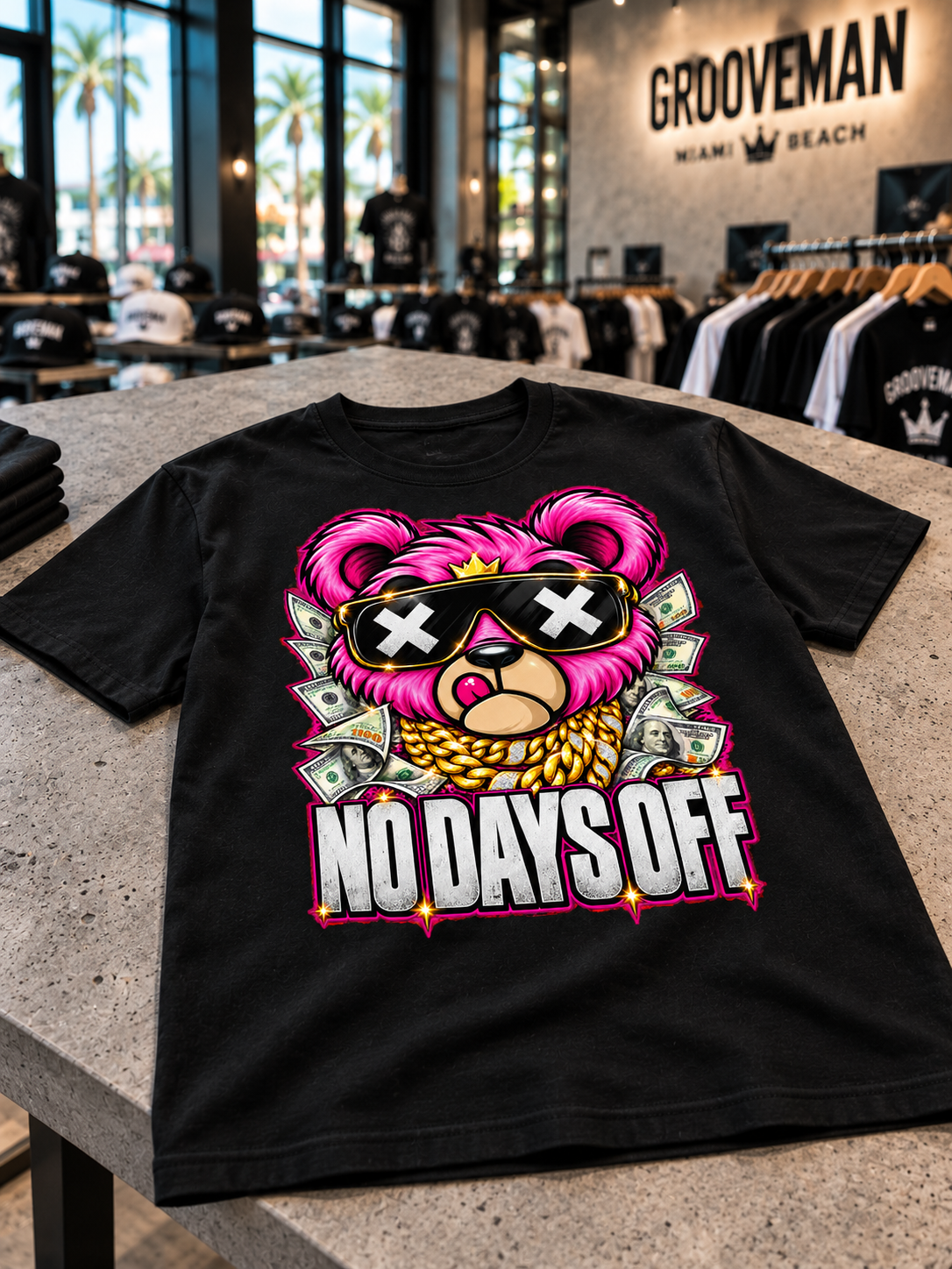 Black No Days Off T-shirt on a concrete display table, featuring a pink crowned teddy, gold-chain artwork and dollar bills.