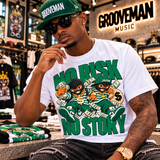 A man wearing a white t-shirt with a green and black 'NO RISK NO STORY' cartoon graphic, sitting in a music store. The t-shirt features a bold green and black design with a cartoon character wearing a green cap and sunglasses, surrounded by money bags.
