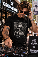 A man wearing a black t-shirt with a large white skull and crown graphic, standing behind a Pioneer DJ mixer in an outdoor street setting with Grooveman Miami Beach signage.