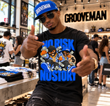 A man wearing a black graphic t-shirt with a blue 'GROOVEMAN' cap and sunglasses, posing inside a retail store. The t-shirt features a cartoon graphic of a character wearing blue sneakers and holding cash, with the text 'NO RISK NO STORY'.