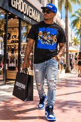 A man walking on a sunny street wearing a black Grooveman t-shirt with a colorful cartoon graphic, light-wash ripped jeans, blue and white sneakers, and a blue cap. He is holding a black Grooveman shopping bag.
