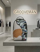 A Grooveman x Sprayground Street backpack displayed on a concrete pedestal inside a modern boutique. The backpack features a split design: one half is a gray and white checkerboard pattern with a tan shark-mouth graphic, and the other half is covered in a stack of dollar bills.