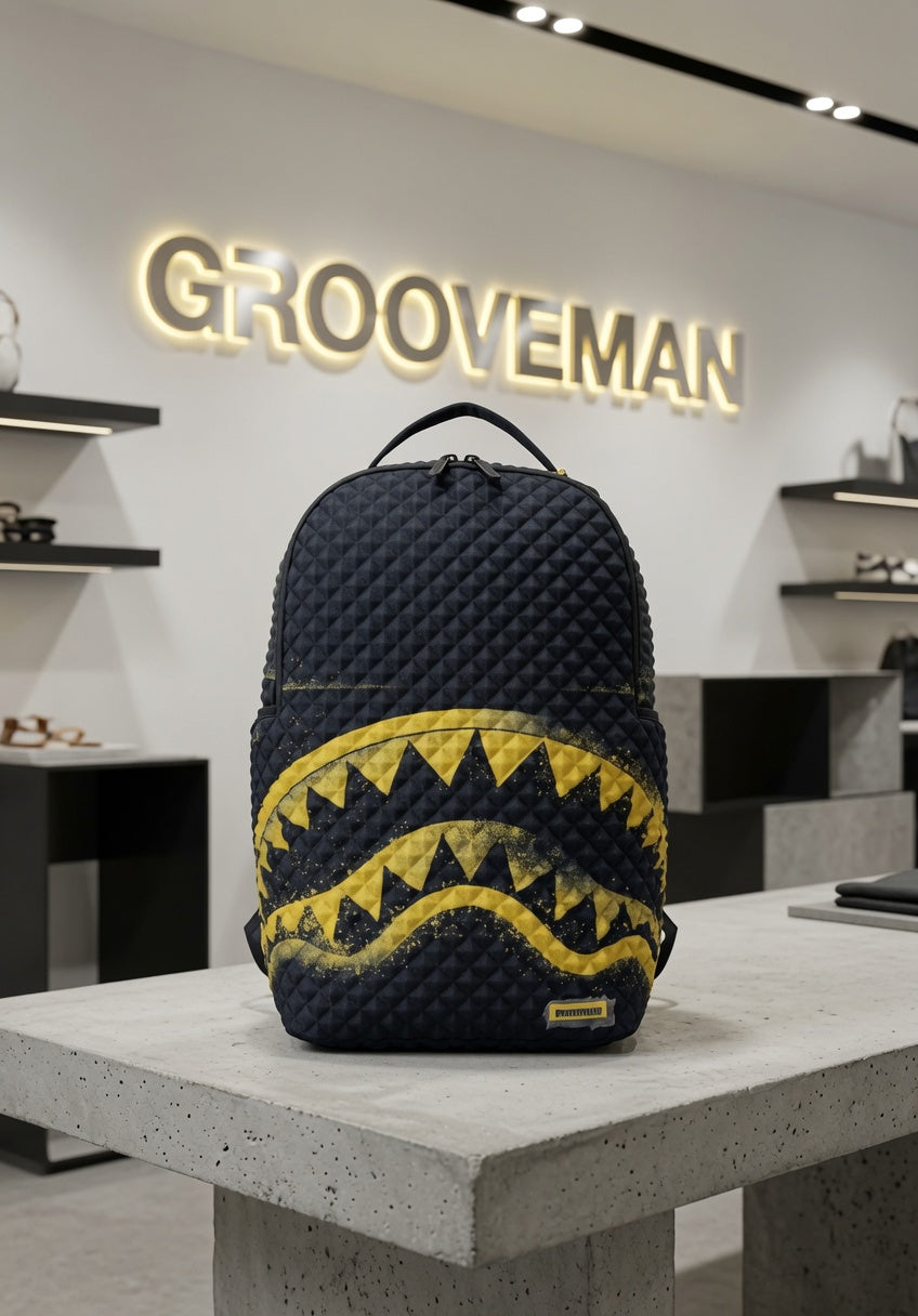 A dark navy blue quilted backpack with a yellow shark-mouth graphic, displayed on a concrete table inside a modern retail store with a GROOVEMAN sign in the background.