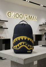 A dark navy blue quilted backpack with a yellow shark-mouth graphic, displayed on a concrete table inside a modern retail store with a GROOVEMAN sign in the background.