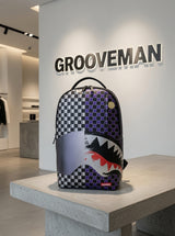 A Grooveman backpack by Sprayground featuring a split checkerboard pattern and a shark mouth graphic, displayed on a concrete pedestal in a minimalist retail store.