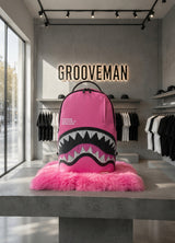 A bright pink Sprayground backpack with a shark mouth graphic, displayed on a pink faux fur rug on a concrete table inside a modern clothing store.