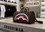 A Grooveman duffle bag with a brown checkerboard pattern and a large shark mouth graphic on the front, displayed on a concrete counter inside a retail store.