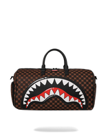 A brown checkered duffel bag featuring a large shark mouth graphic on the front with white teeth, a red interior, and black outlines. The bag has black top handles and is shown against a plain white background.
