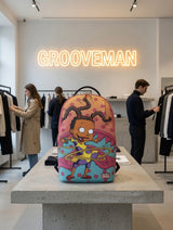 A colorful Sprayground backpack featuring a cartoon character design, displayed on a concrete table inside a clothing store with a glowing Grooveman neon sign in the background.