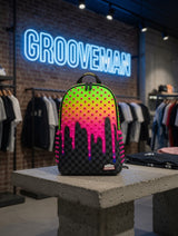 A black and neon gradient backpack with a dripping city skyline design sits on a concrete display table inside a retail store. The background features a brick wall with a glowing blue 'GROOVEMAN' neon sign and clothing racks.