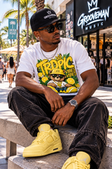 A man sitting on a concrete bench outdoors wearing a white graphic t-shirt with a 'Tropic Vibes' cartoon bear design, a black cap with a white logo, dark washed jeans, and yellow sneakers. The background shows a sunny street scene with palm trees and a 'Groovemusic' storefront.