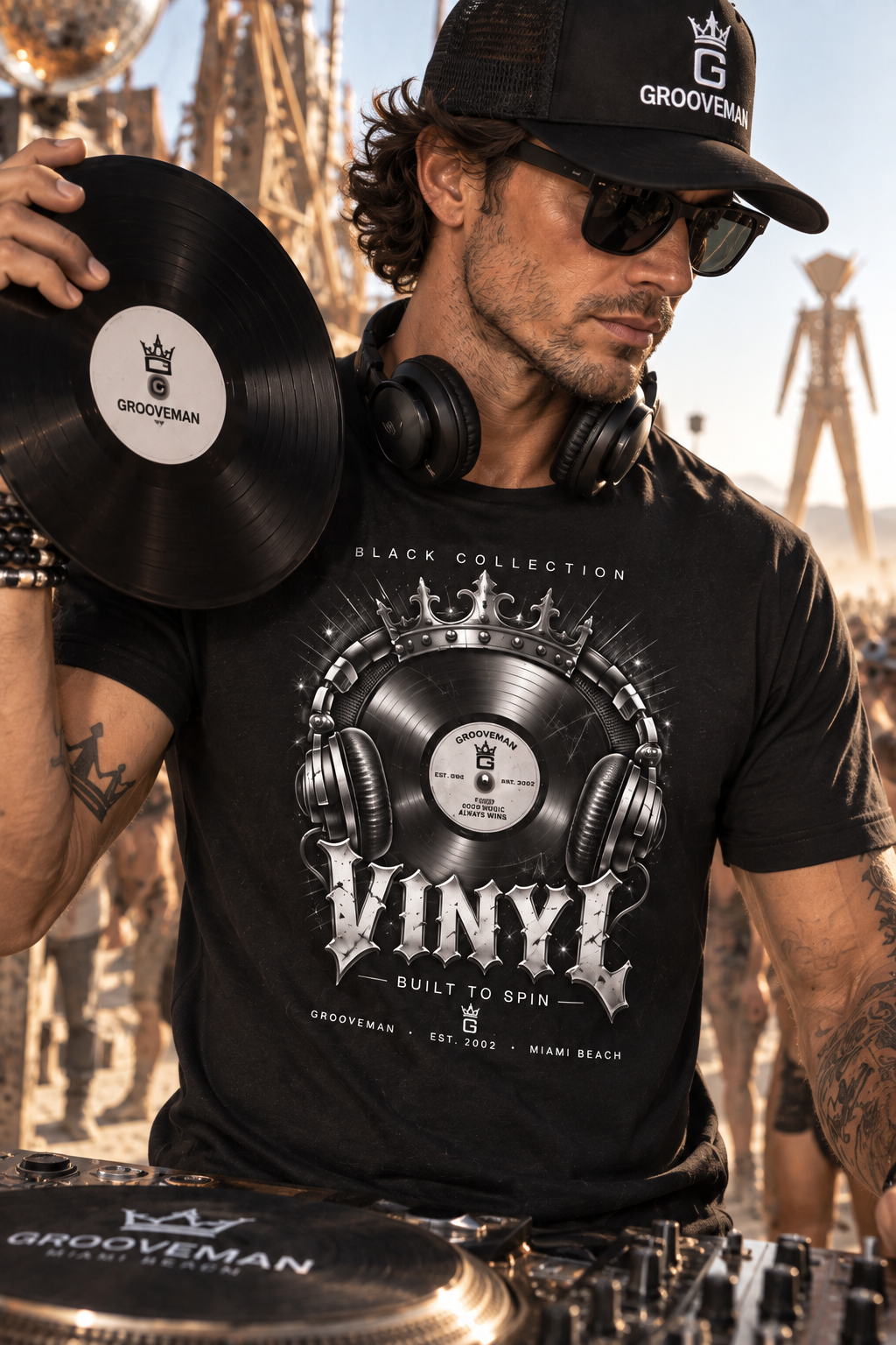 A man wearing a black Grooveman t-shirt and matching black cap, holding a vinyl record in front of a DJ setup with a blurred outdoor background.