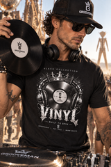 A man wearing a black Grooveman t-shirt and matching black cap, holding a vinyl record in front of a DJ setup with a blurred outdoor background.